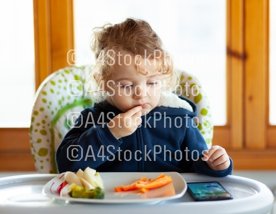 Toddler eats while watching movies on the mobile phone.